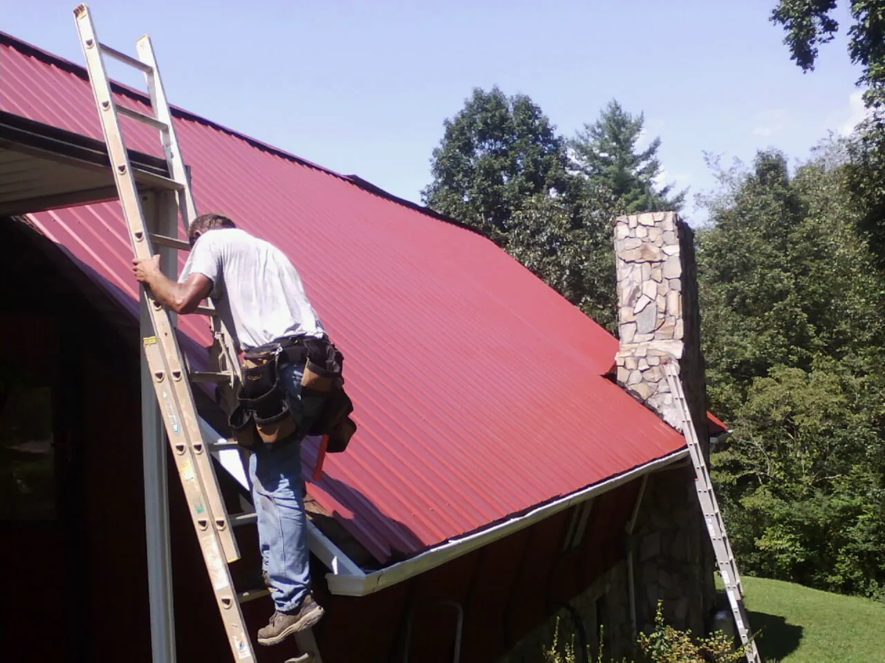 Roofer on a ladder beside a red metal roof and stone chimney — Soffit & Fascia Repair by Roofing Brothers in De Soto